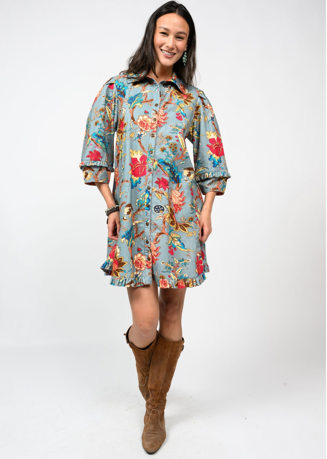 Woman wearing a medium blue dress with a multicolor floral pattern and brown boots in front of a white background