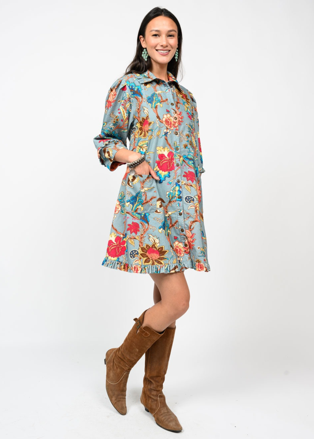 Woman wearing a medium blue dress with a multicolor floral pattern and brown boots in front of a white background