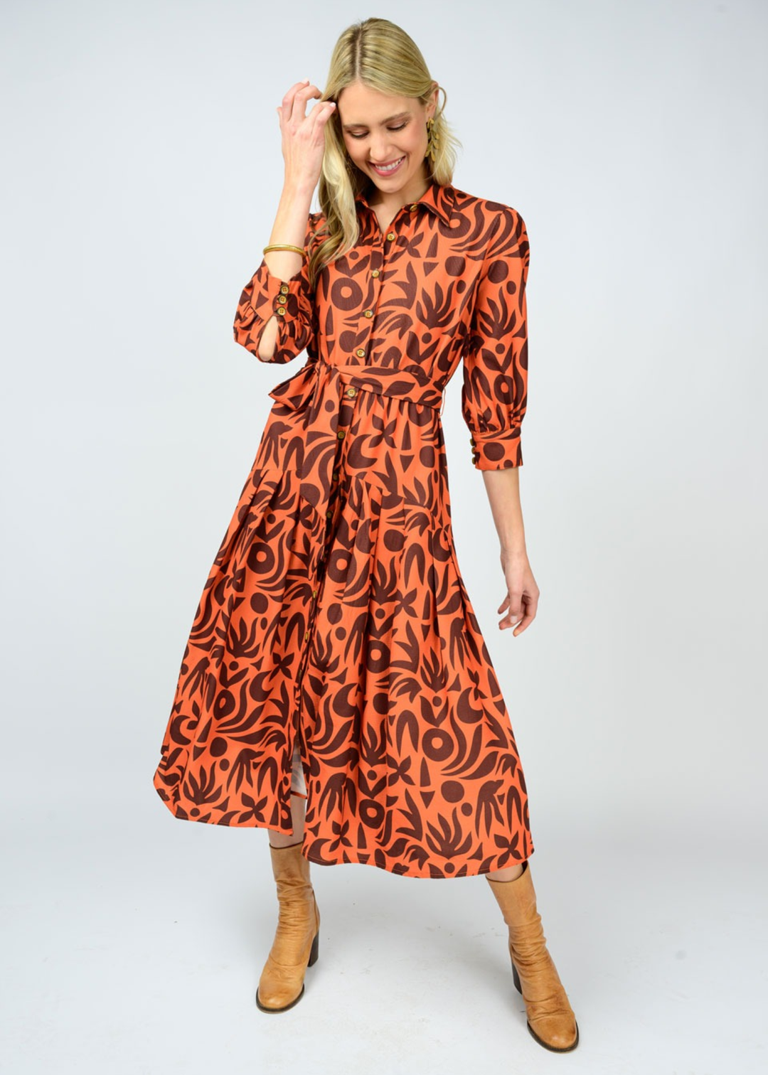 Woman holding hair wearing an orange and brown patterned midi dress with light brown boots in front of a white background