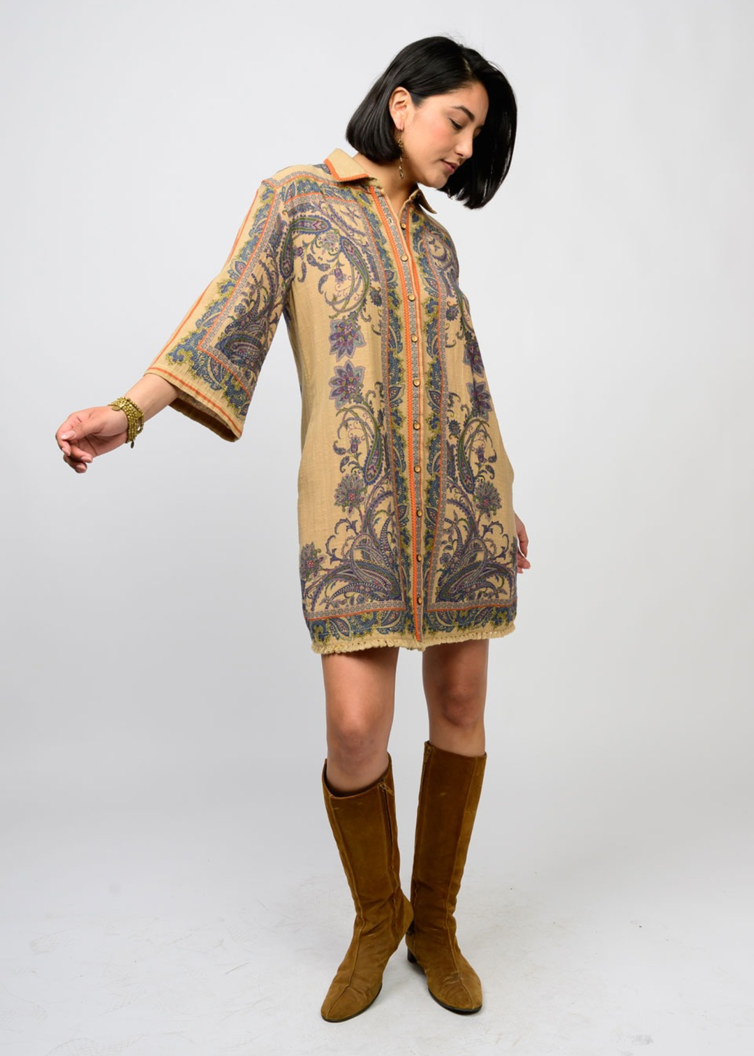 Woman wearing a camel and floral button down dress with 3/4 length sleeves and tall brown boots standing in front of a white background