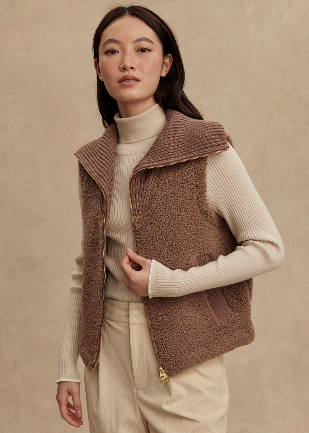 Woman wearing a brown vest over a cream turtleneck and cream pants standing in front of a tan background