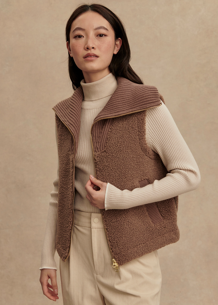 Woman wearing a brown vest over a cream turtleneck and cream pants standing in front of a tan background