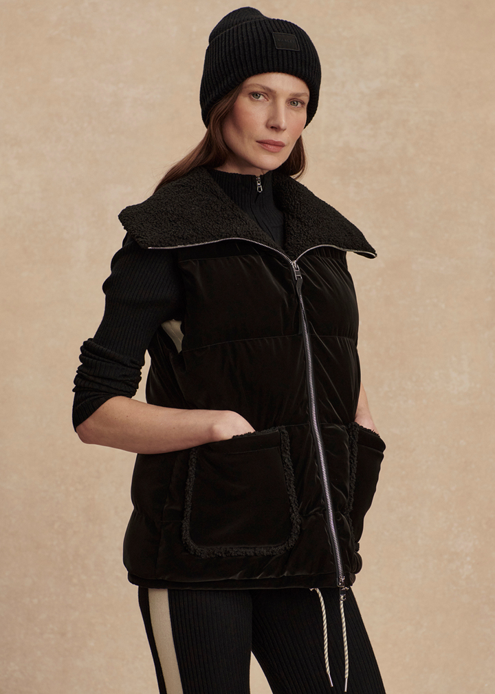 Woman with hand in pocket wearing a black outfit with a black puffer vest against a tan background