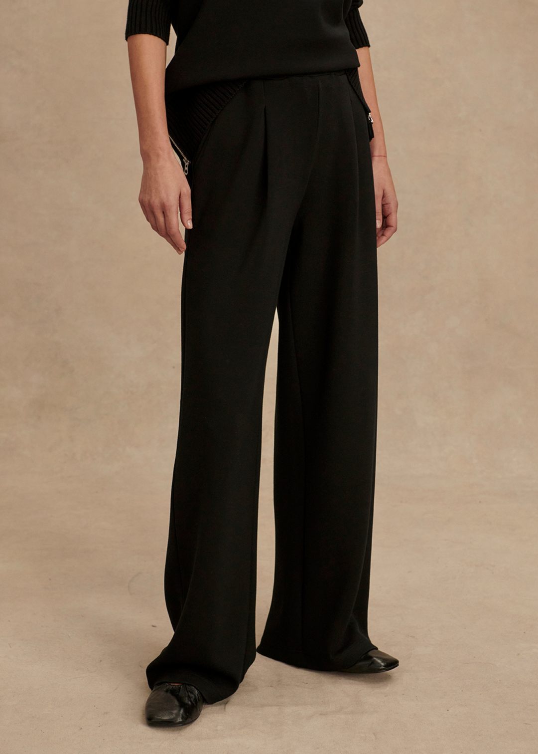 Woman wearing long black wide leg sweatpants with a black top in front of a tan background