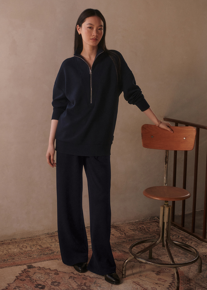 Woman leaning against a chair wearing wide leg navy sweatpants with a matching top in front of a tan background