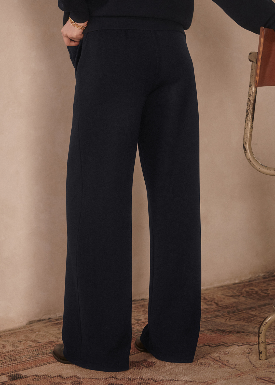 Close up of a woman turned around wearing wide leg navy sweatpants with a matching top in front of a tan background