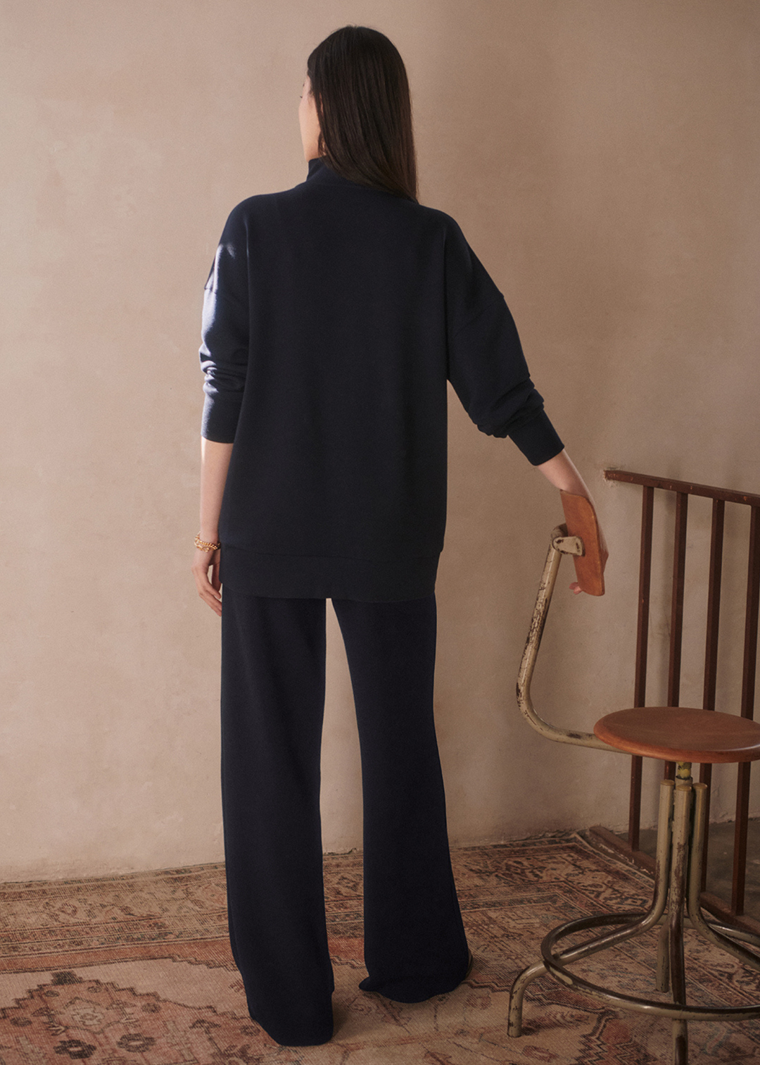 Woman turned around wearing wide leg navy sweatpants with a matching top in front of a tan background