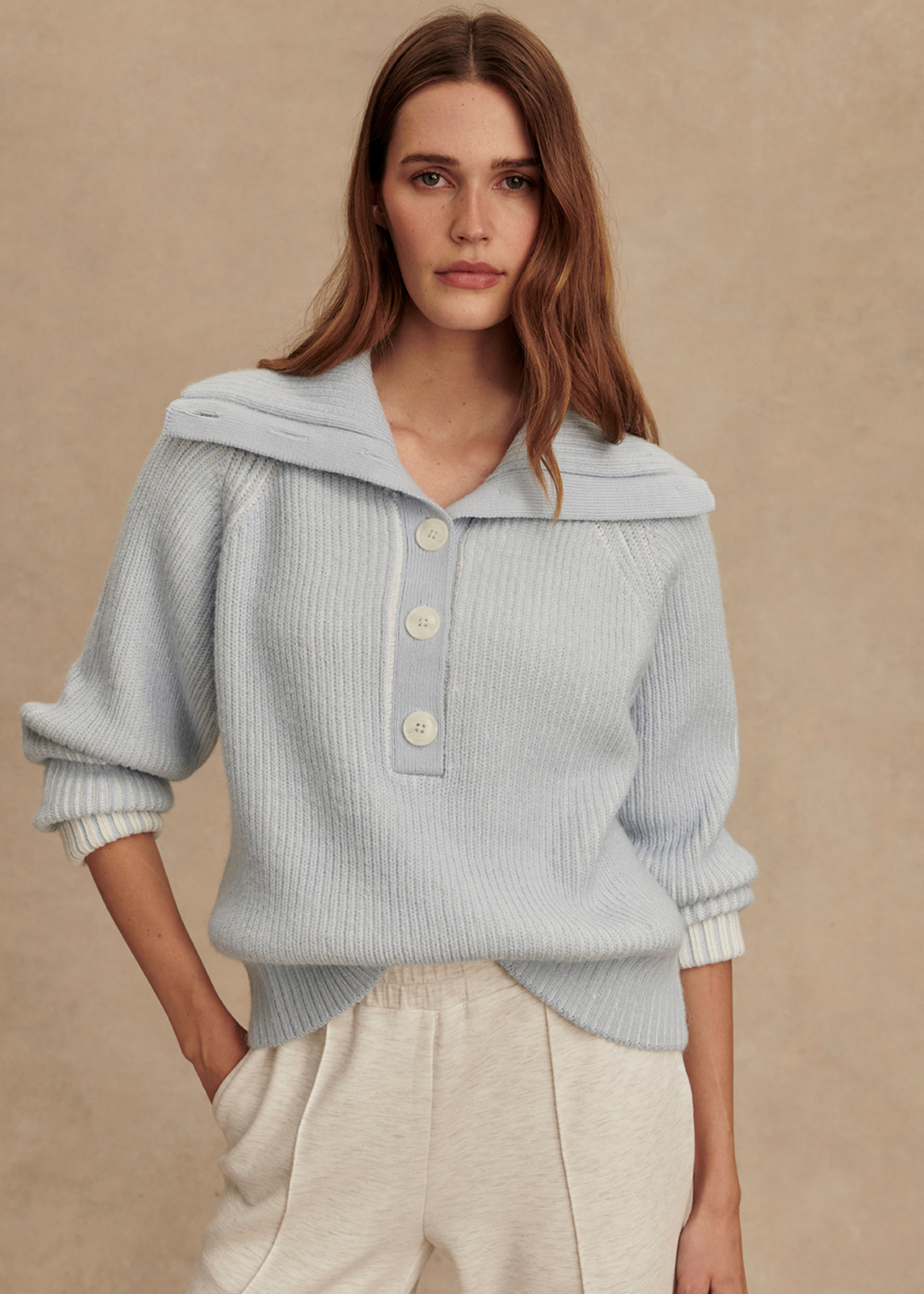 Woman wearing a light blue ribbed sweater with white pants in front of a tan background with hand in pocket