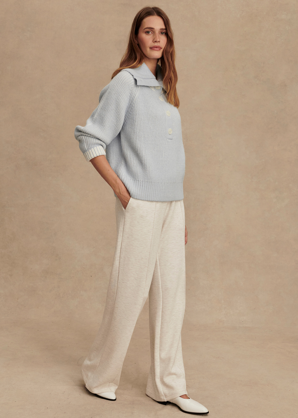 Woman turned to the side wearing a light blue ribbed sweater with white pants in front of a tan background with hand in pocket