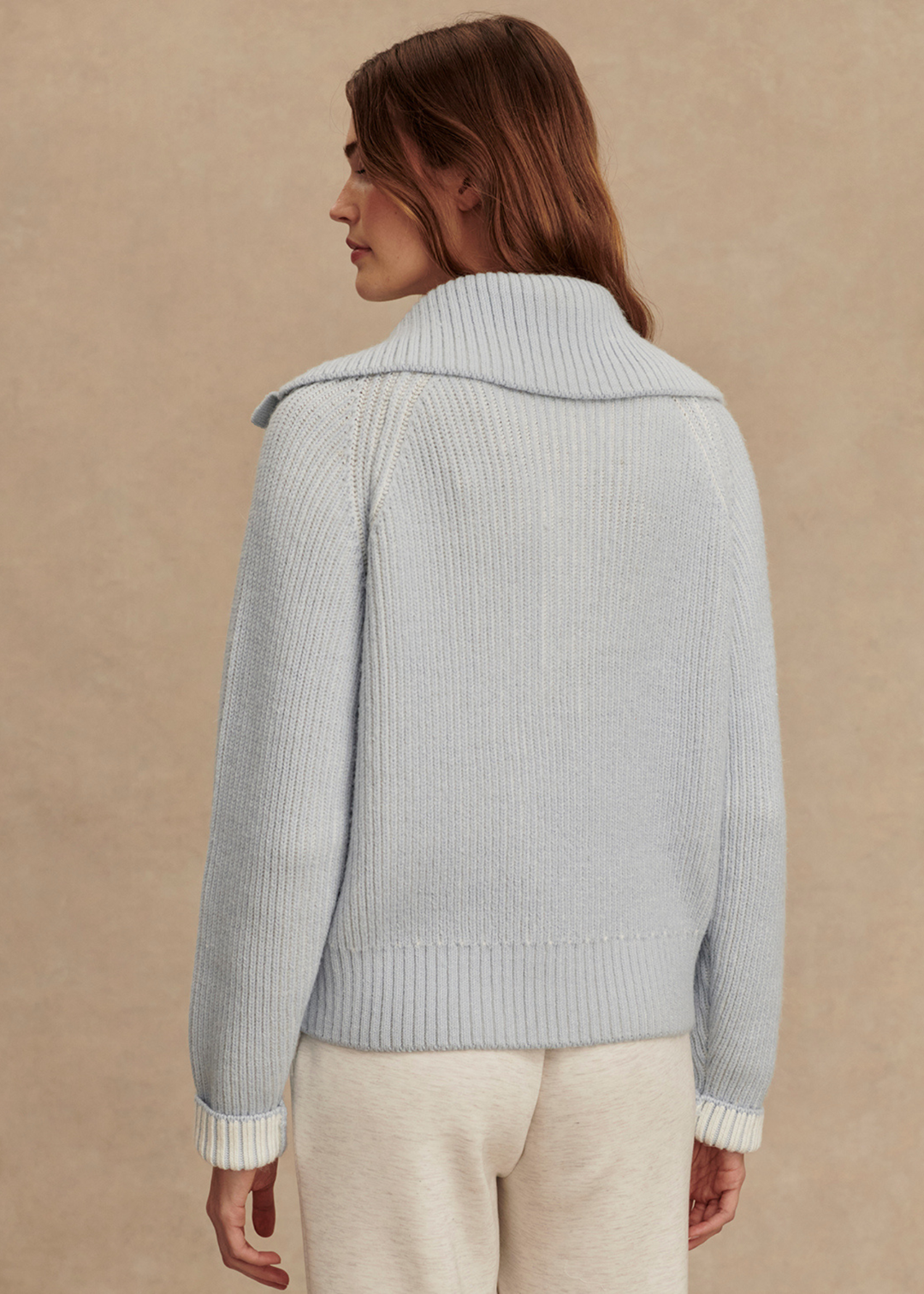 Woman turned around wearing a light blue ribbed sweater with white pants in front of a tan background