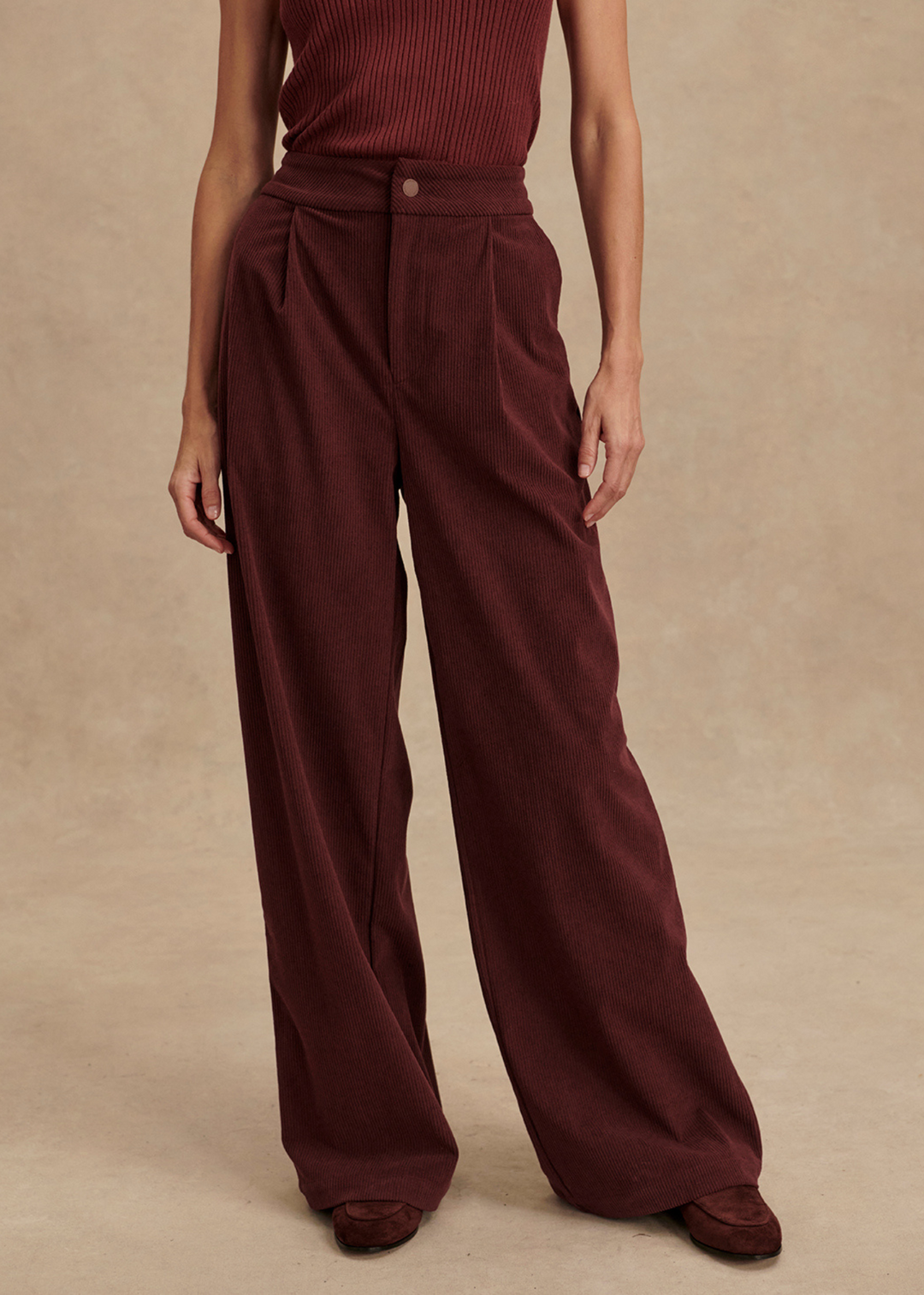 Woman wearing brown corduroy pants with a red tank in front of a tan background