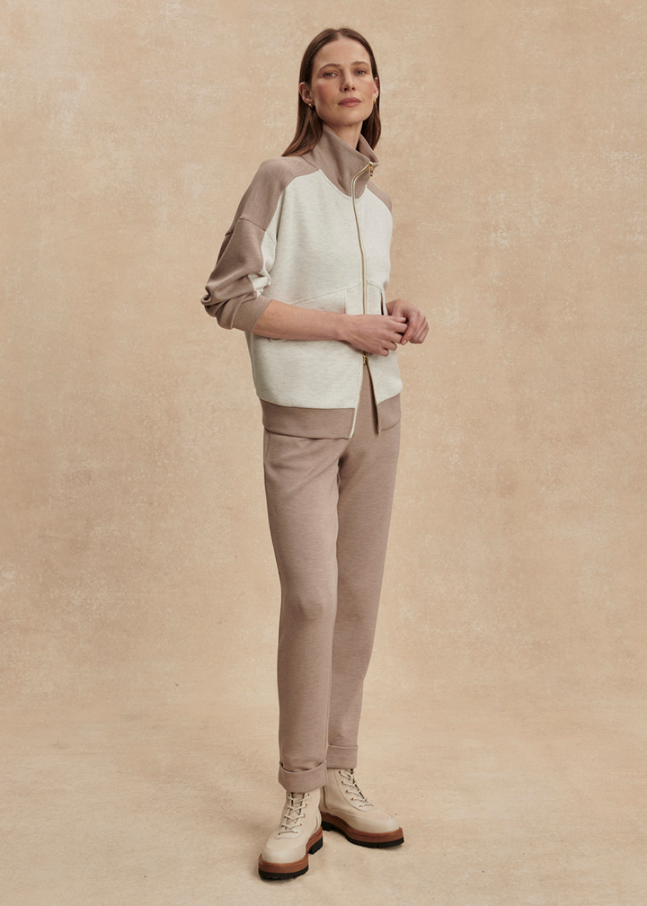 Woman wearing a taupe and white zip up sweatshirt with taupe pants against a tan background
