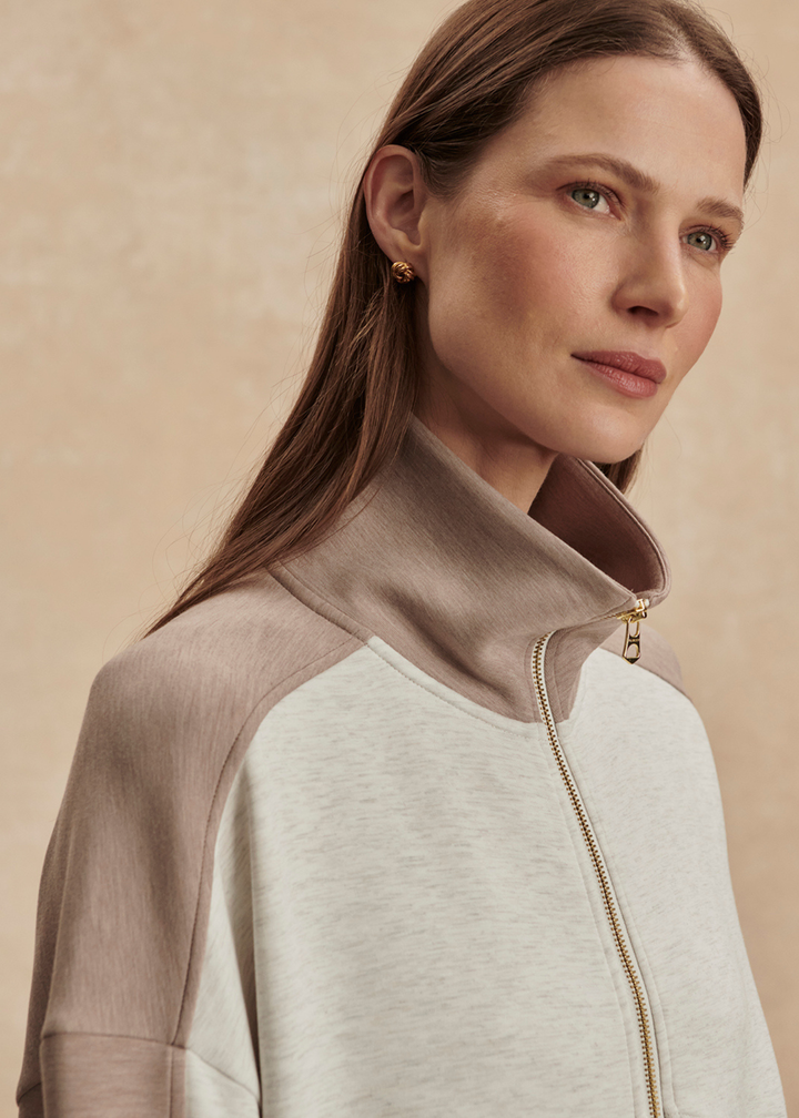 Close up of a woman wearing a taupe and white zip up sweatshirt against a tan background