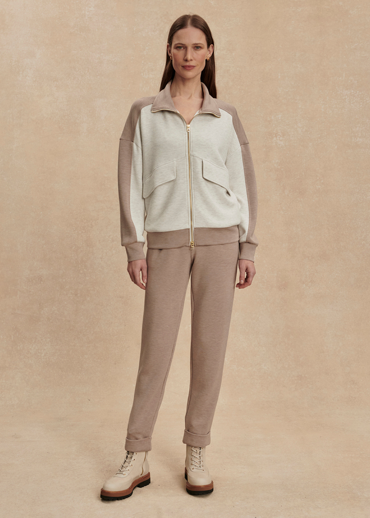 Woman wearing a taupe and white zip up sweatshirt with taupe pants against a tan background