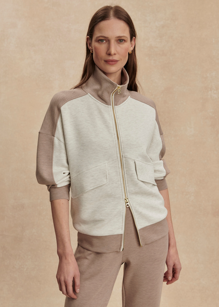 Woman wearing a taupe and white zip up sweatshirt with taupe pants against a tan background