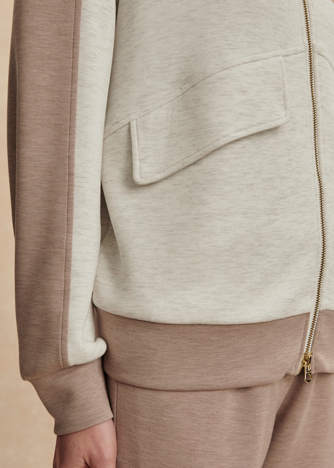 Close up of a woman wearing a taupe and white zip up sweatshirt with taupe pants against a tan background