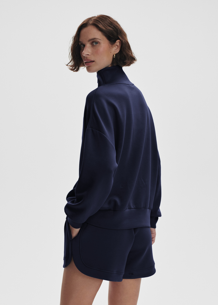 Woman turned to the side wearing a navy half zip sweatshirt top with matching shorts in front of a white background