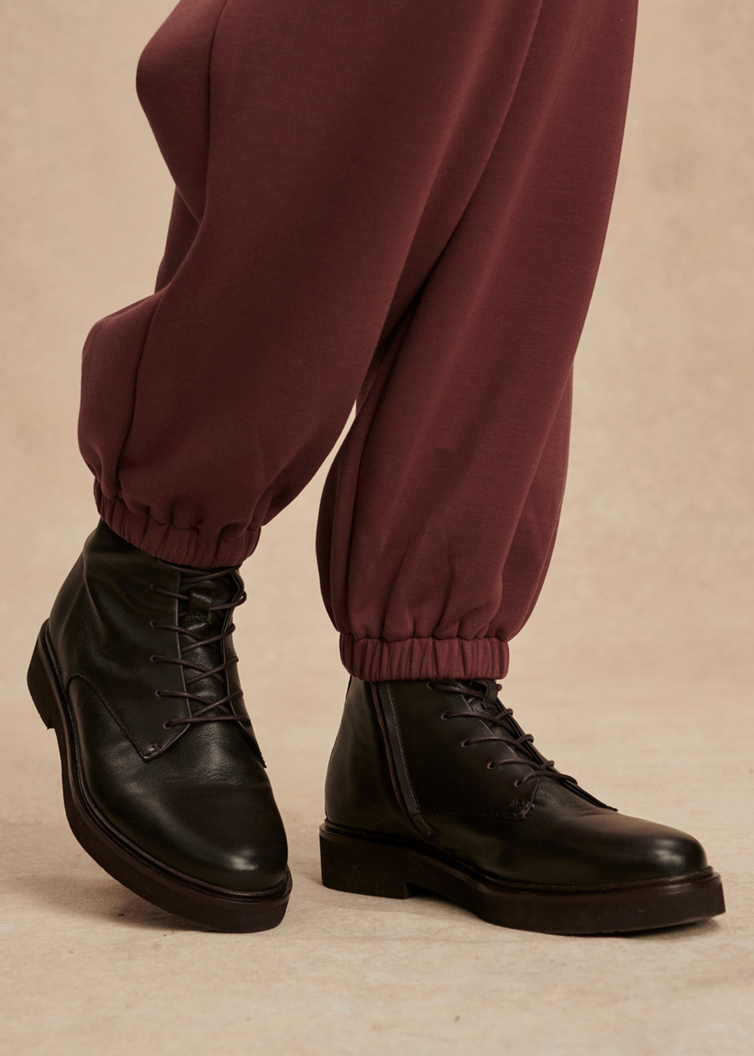 Close up of the ankles of a brown jumpsuit with black boots in front of a tan background