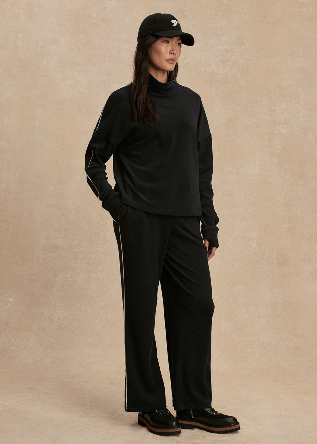 Woman with hand in pocket wearing a black set with white trim against a tan background