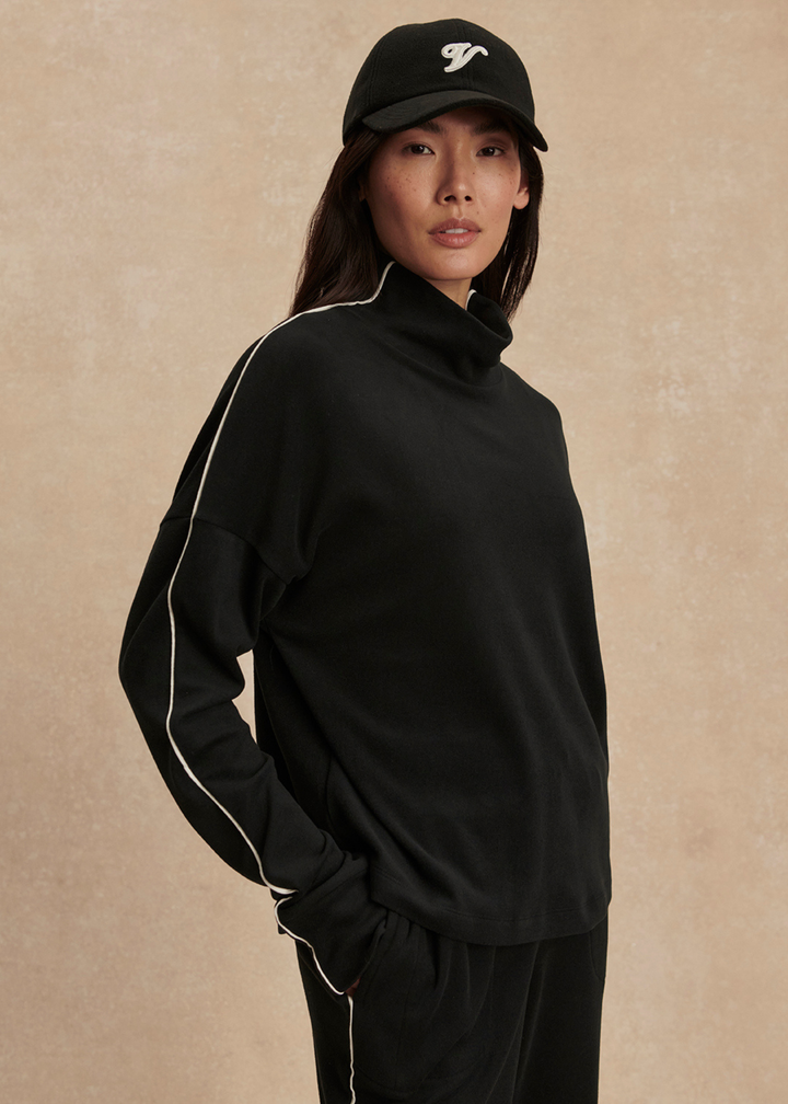 Woman with hand in pocket wearing a black set with white trim against a tan background