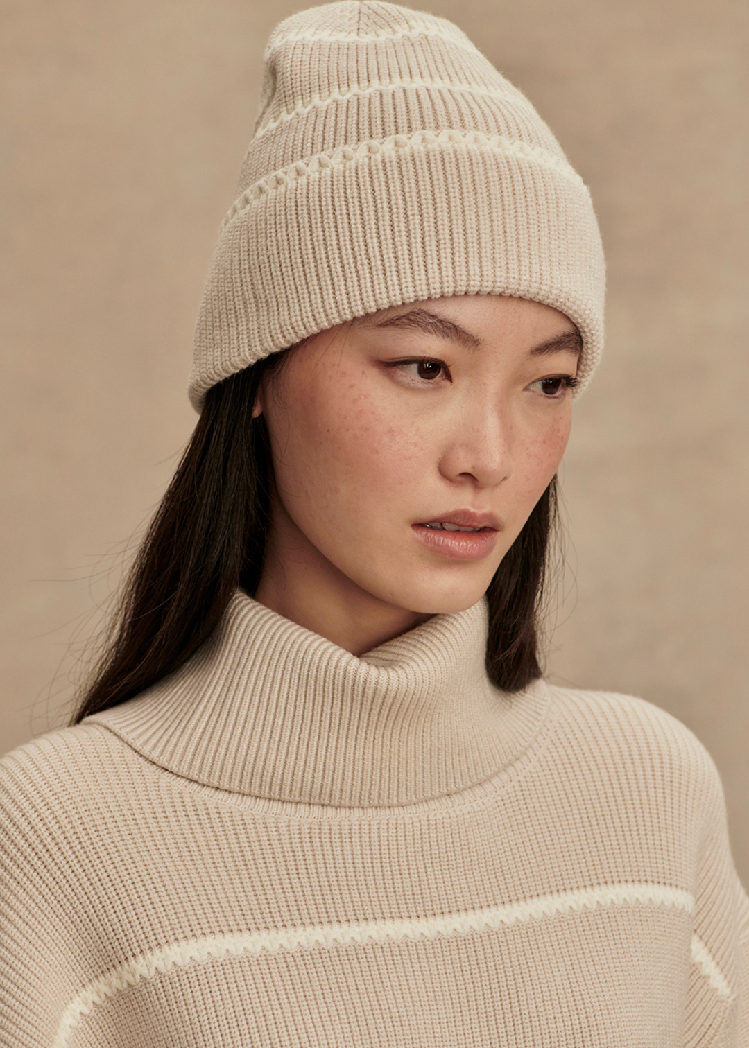 Woman wearing a tan and white striped beanie with a matching sweater in front of a tan background