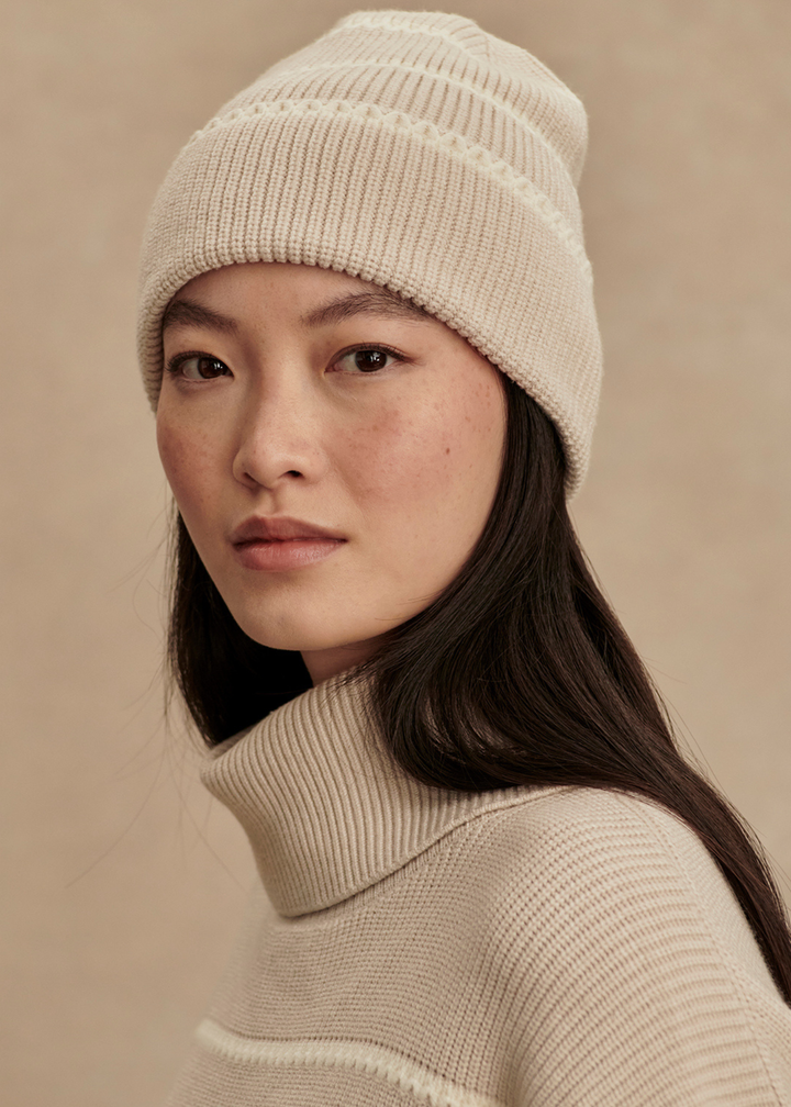 Woman wearing a tan and white striped beanie with a matching sweater in front of a tan background