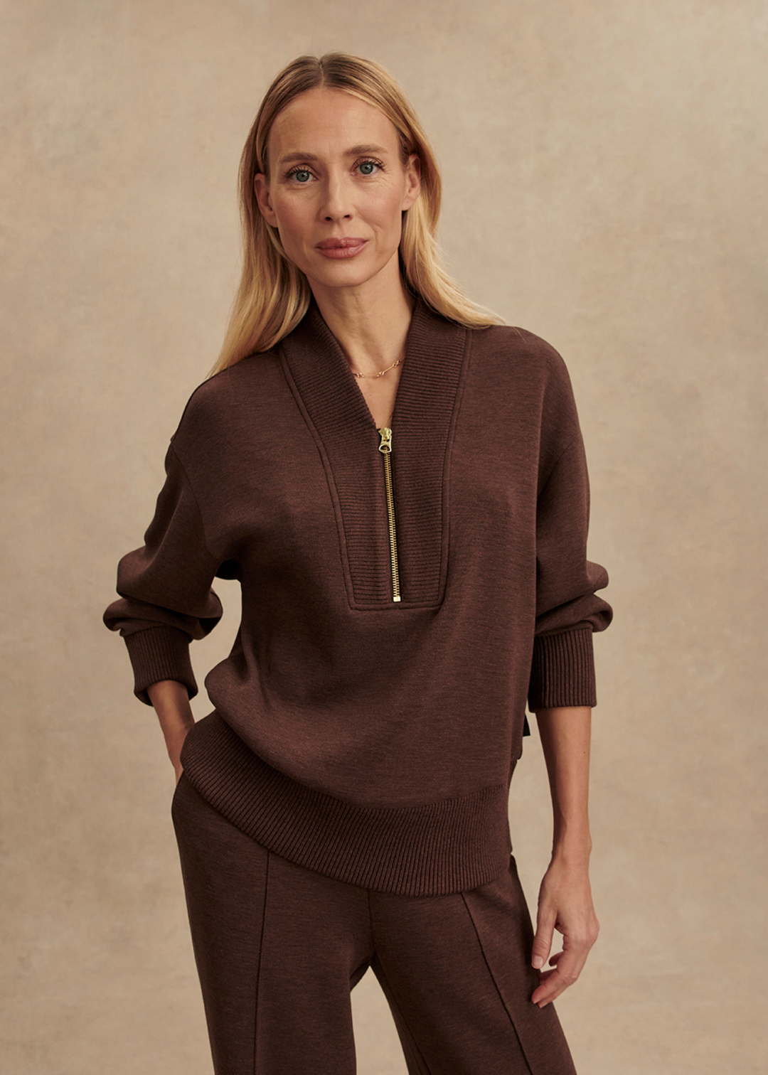 Woman wearing a brown half zip sweatshirt with a gold zipper and matching pants in front of a tan background