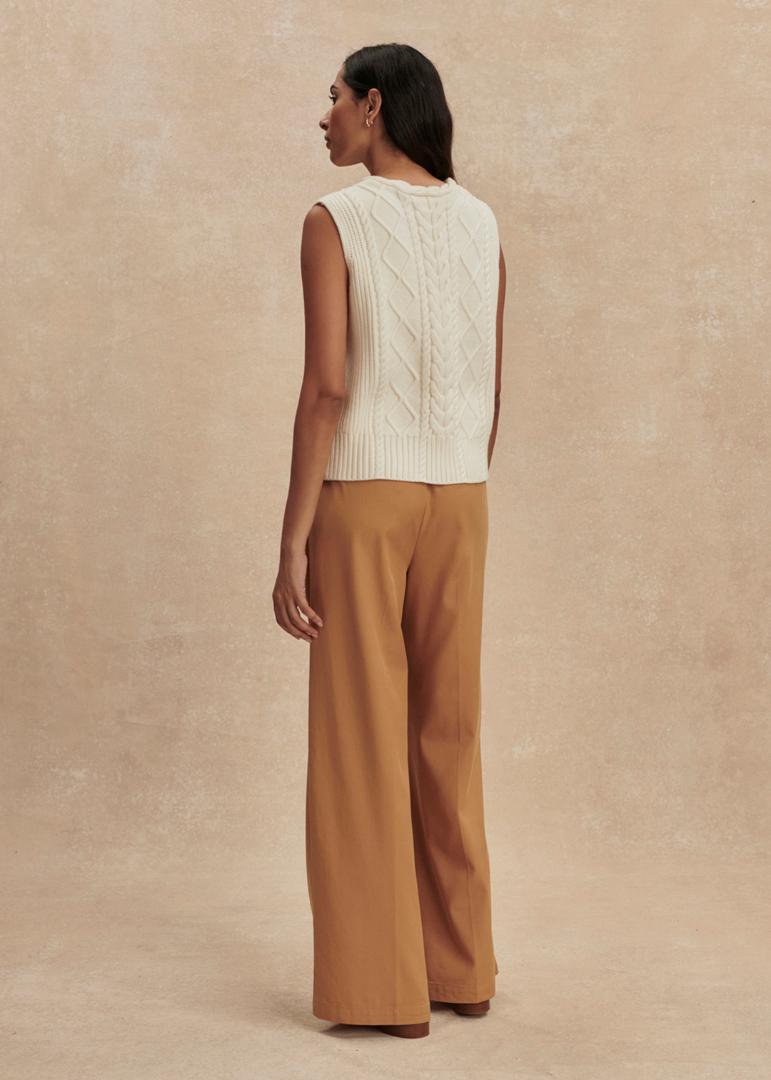 Woman turned around wearing a cream cable knit sweater vest with brown pants