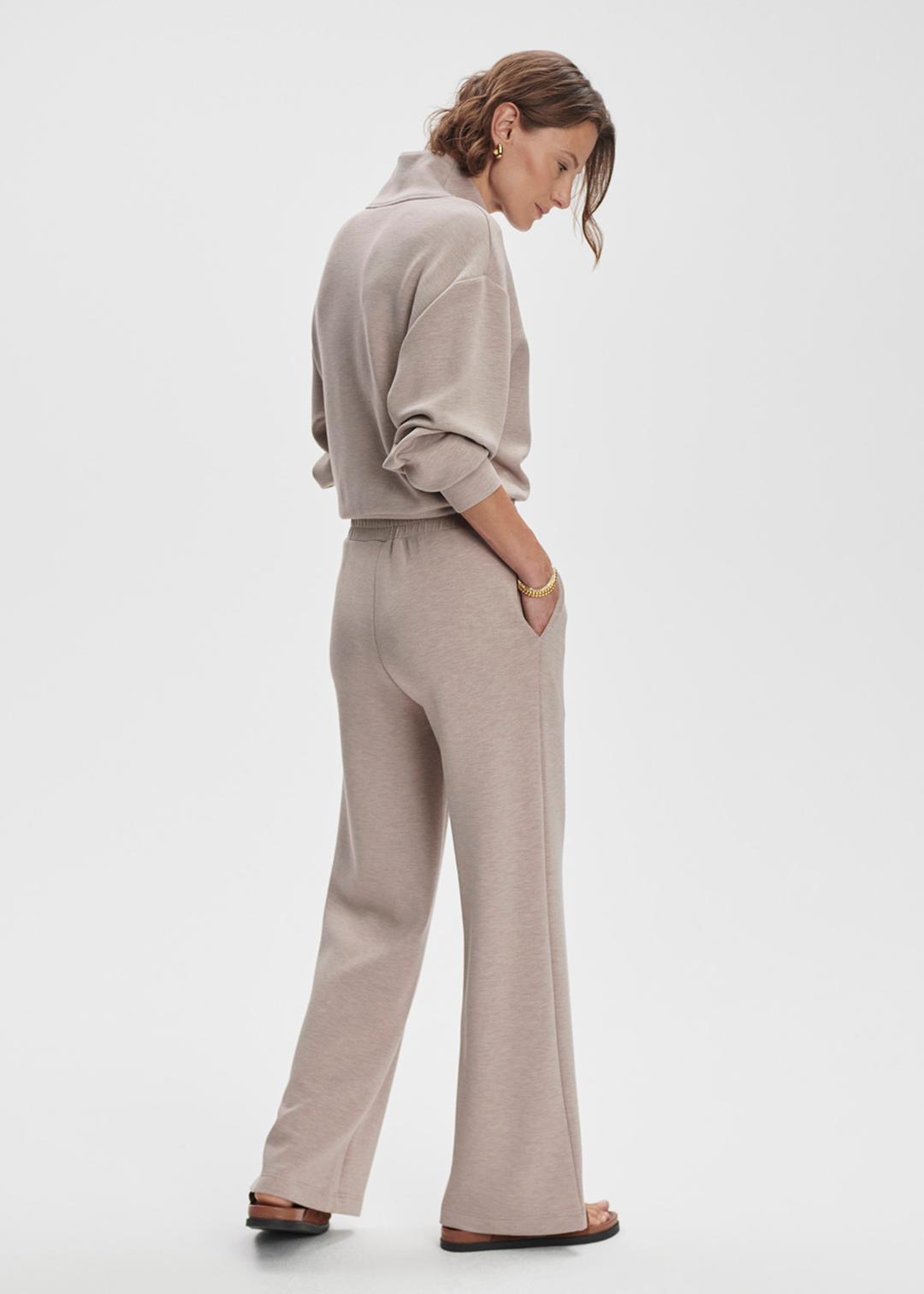 Woman turned to the side wearing taupe wide leg pants with a taupe half zip top against a white background