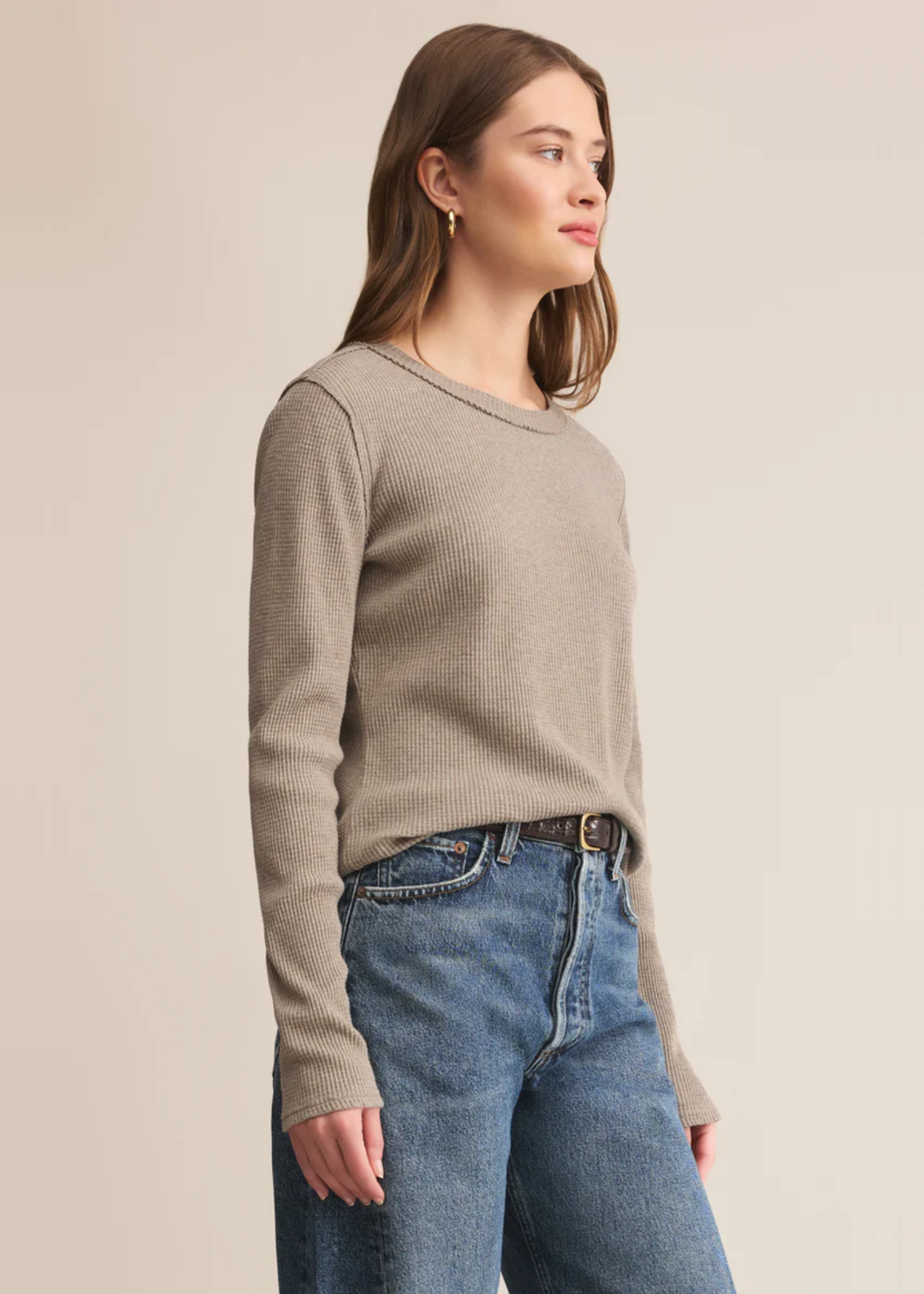 Woman turned to the side wearing a beige colored long sleeve top with jeans against a tan background