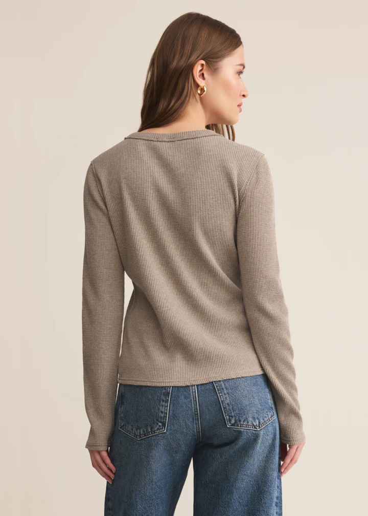 Woman turned around wearing a beige colored long sleeve top with jeans against a tan background