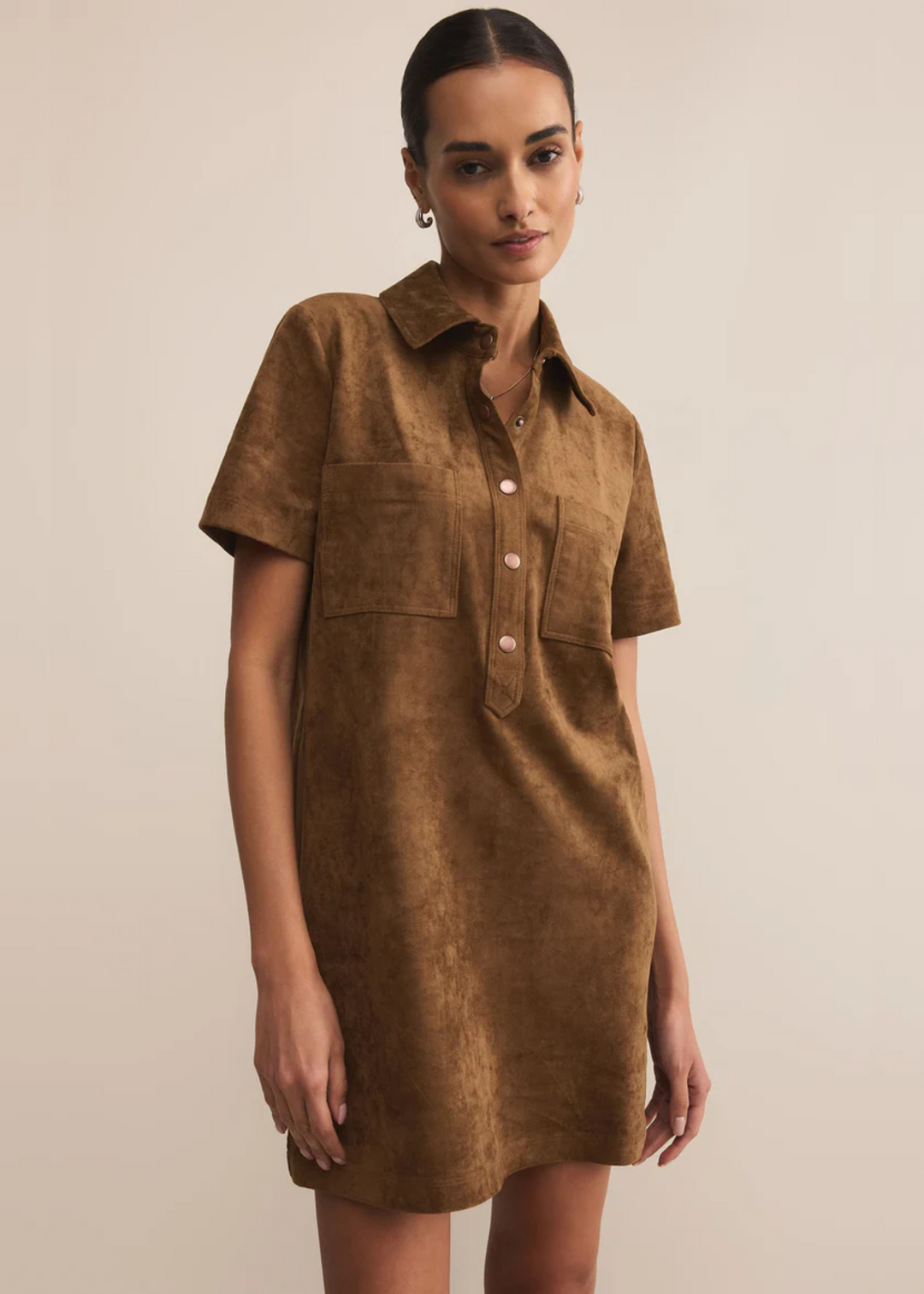 Woman wearing a brown suede dress with a collared neckline and gold button details in front of a grey background