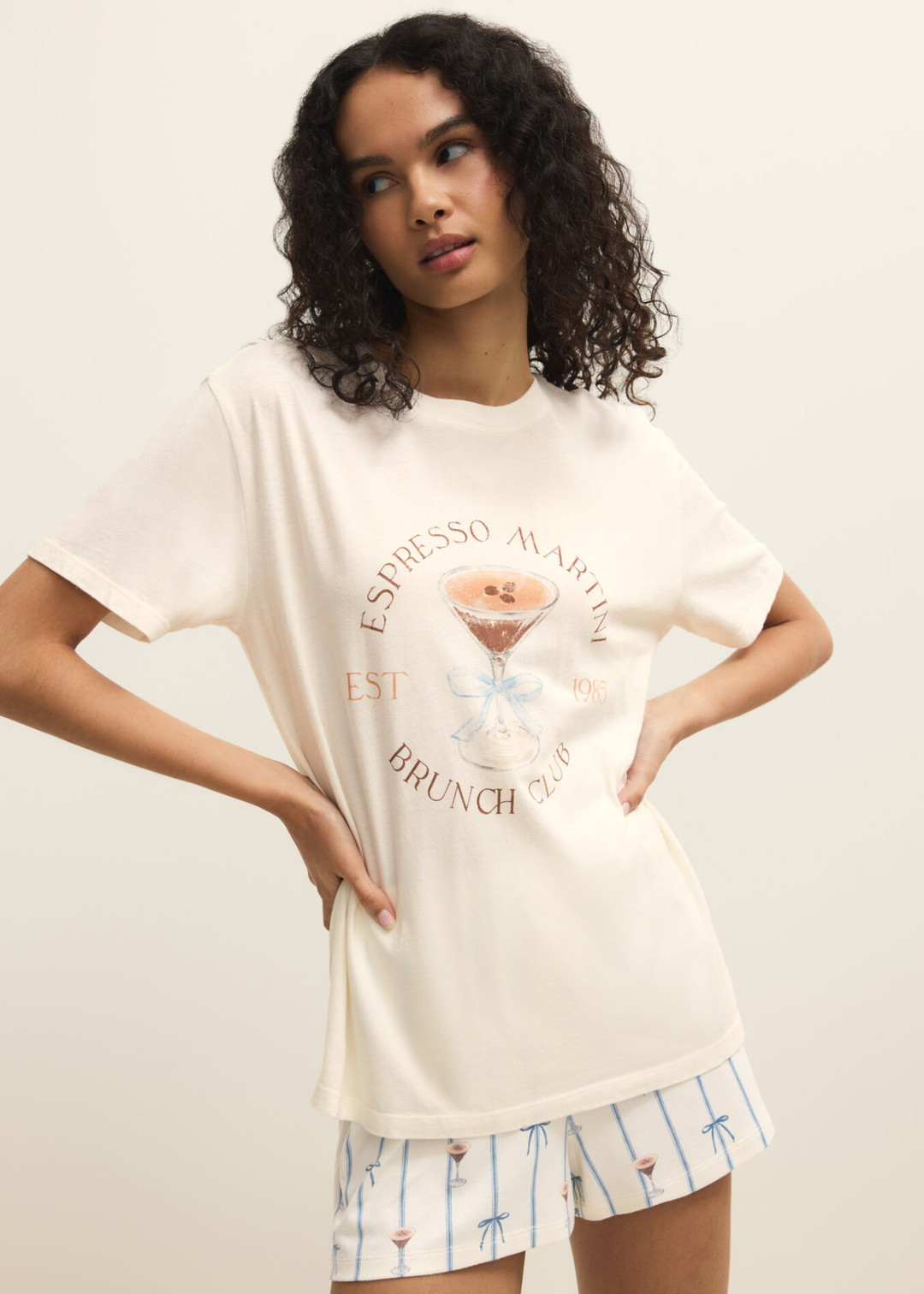 Woman wearing a white shirt with an espresso martini graphic and shorts with hands on hips against a white background