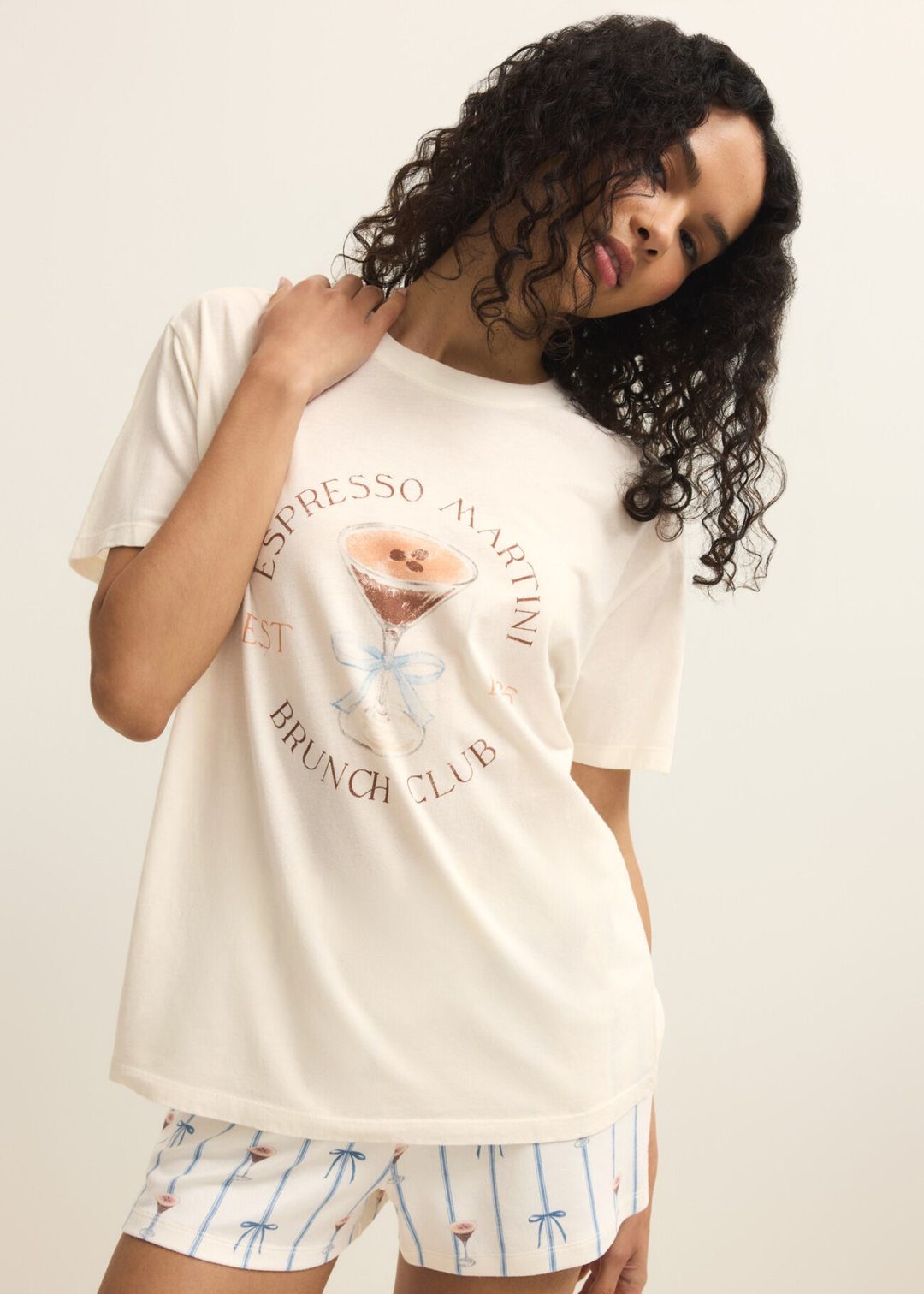 Woman wearing a white shirt with an espresso martini graphic and shorts against a white background