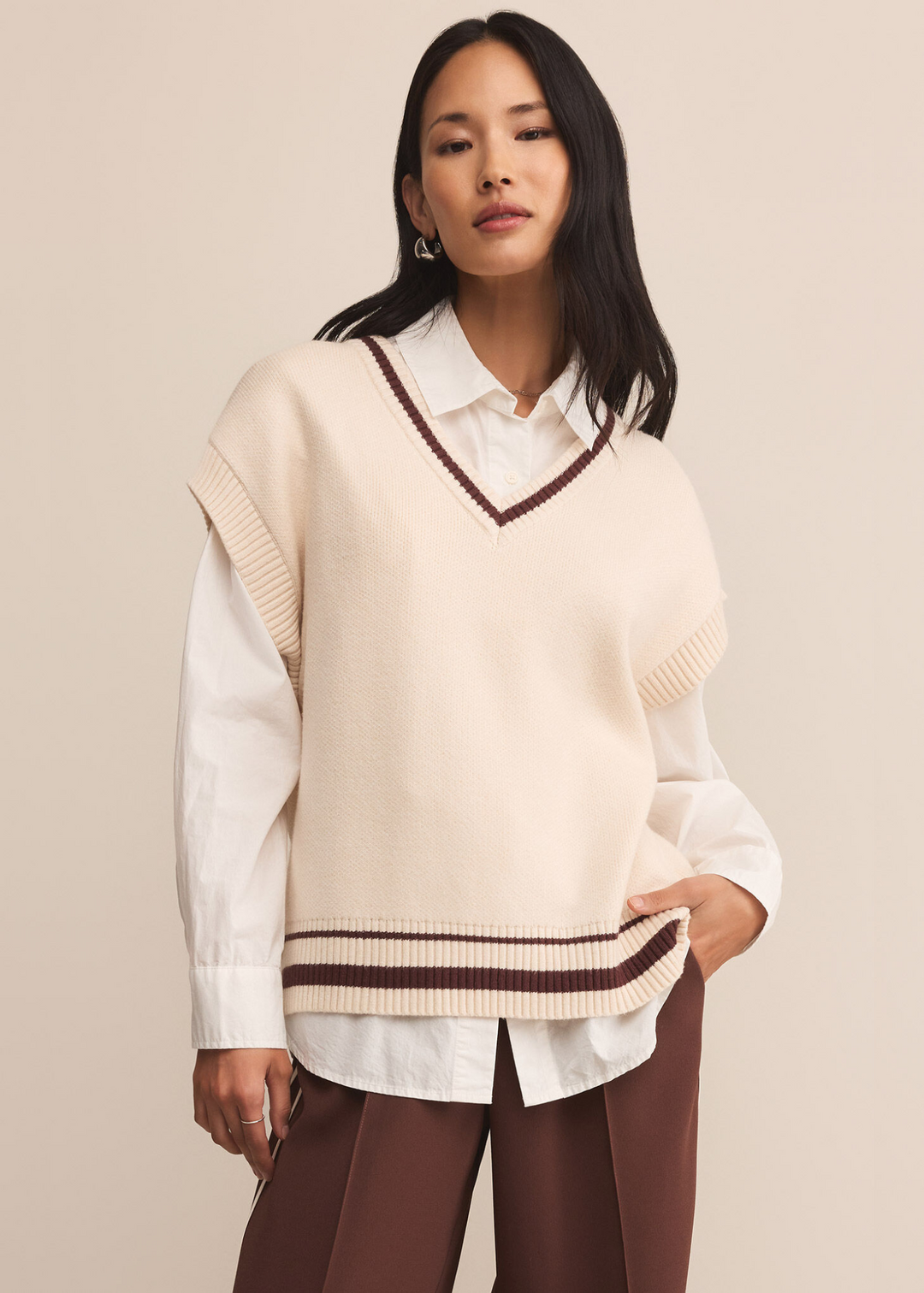 Woman wearing a cream sweater vest with brown stripe trim over a white button down
