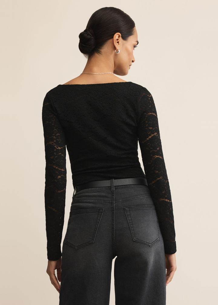 Woman turned around wearing a black lace bodysuit with scalloped trim and black jeans against a tan background