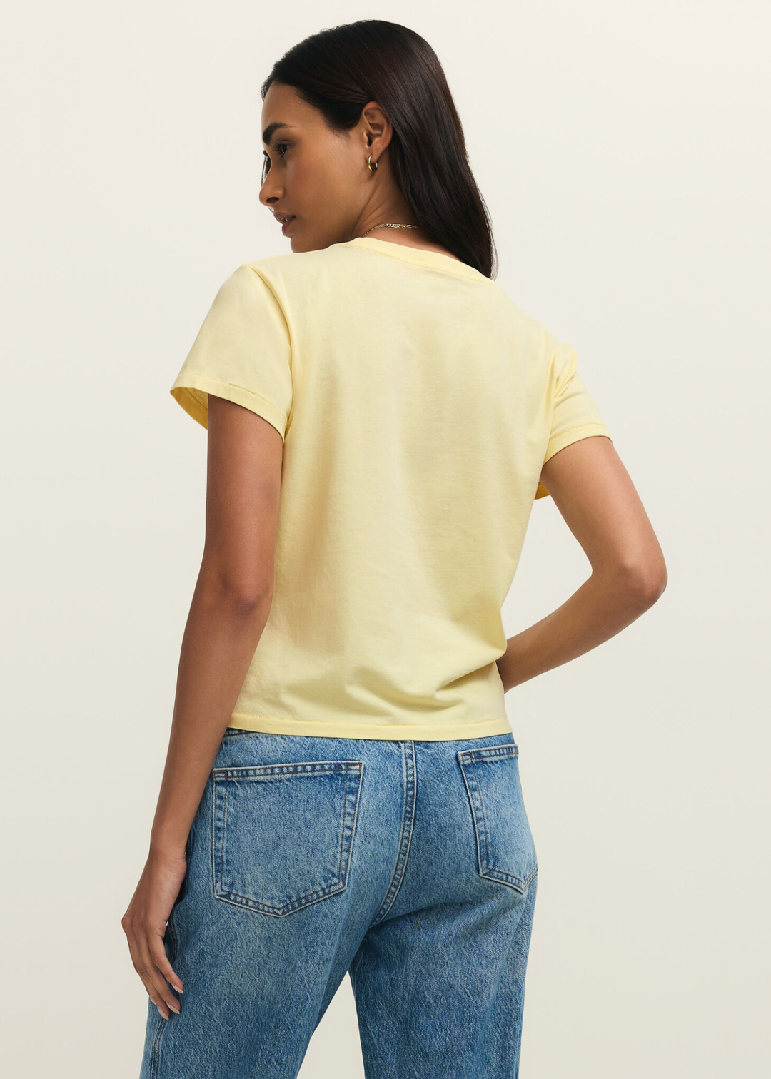 Woman turned around wearing a yellow t-shirt and jeans against a grey background