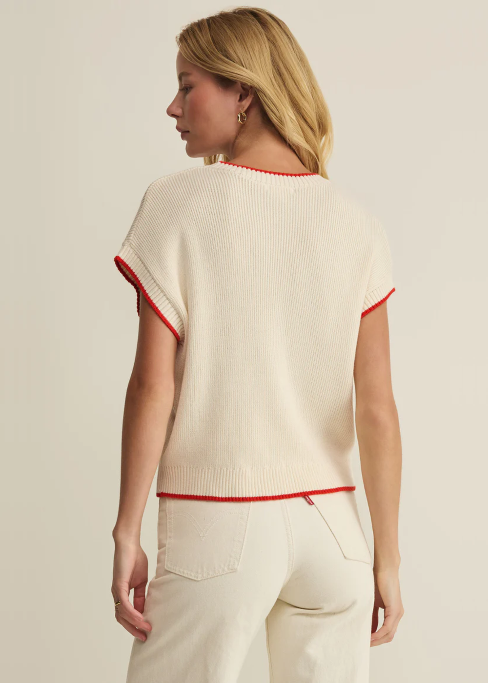 Woman turned around wearing a cream sleeveless sweater with an American flag graphic and white jeans