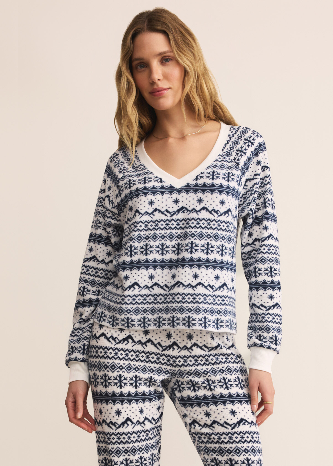 Woman wearing a blue and white fairisle patterned v-neck pajama top with matching pants in front of a tan background