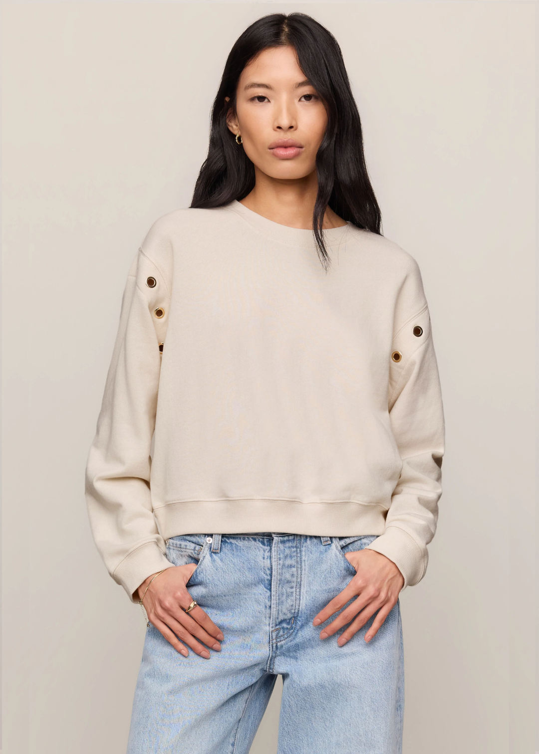 A woman modeling a cream-colored crewneck sweatshirt with decorative gold buttons on the sleeves, paired with light blue jeans, against a solid beige background.