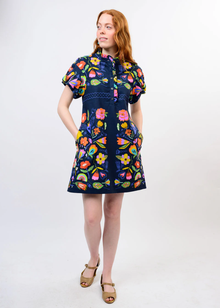 A woman modeling a short-sleeve navy blue dress with a colorful floral print, standing against a plain white studio background.