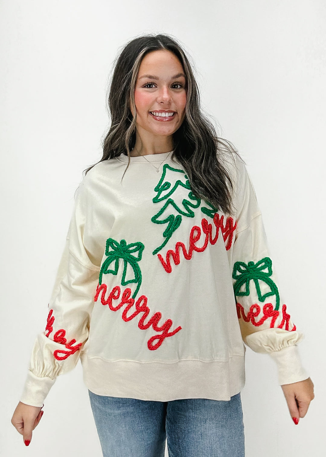 Cream-colored Christmas sweatshirt with festive "Merry" text, holiday bows, and tree accents in red and green.