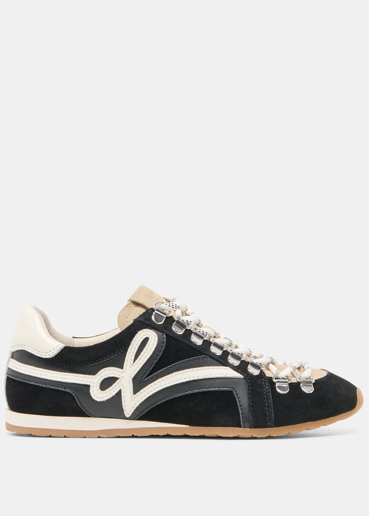 A black suede low-top sneaker with cream-colored panels, a large white script logo on the side, and decorative silver-tone metal eyelets on the laces, shown against a light gray background.