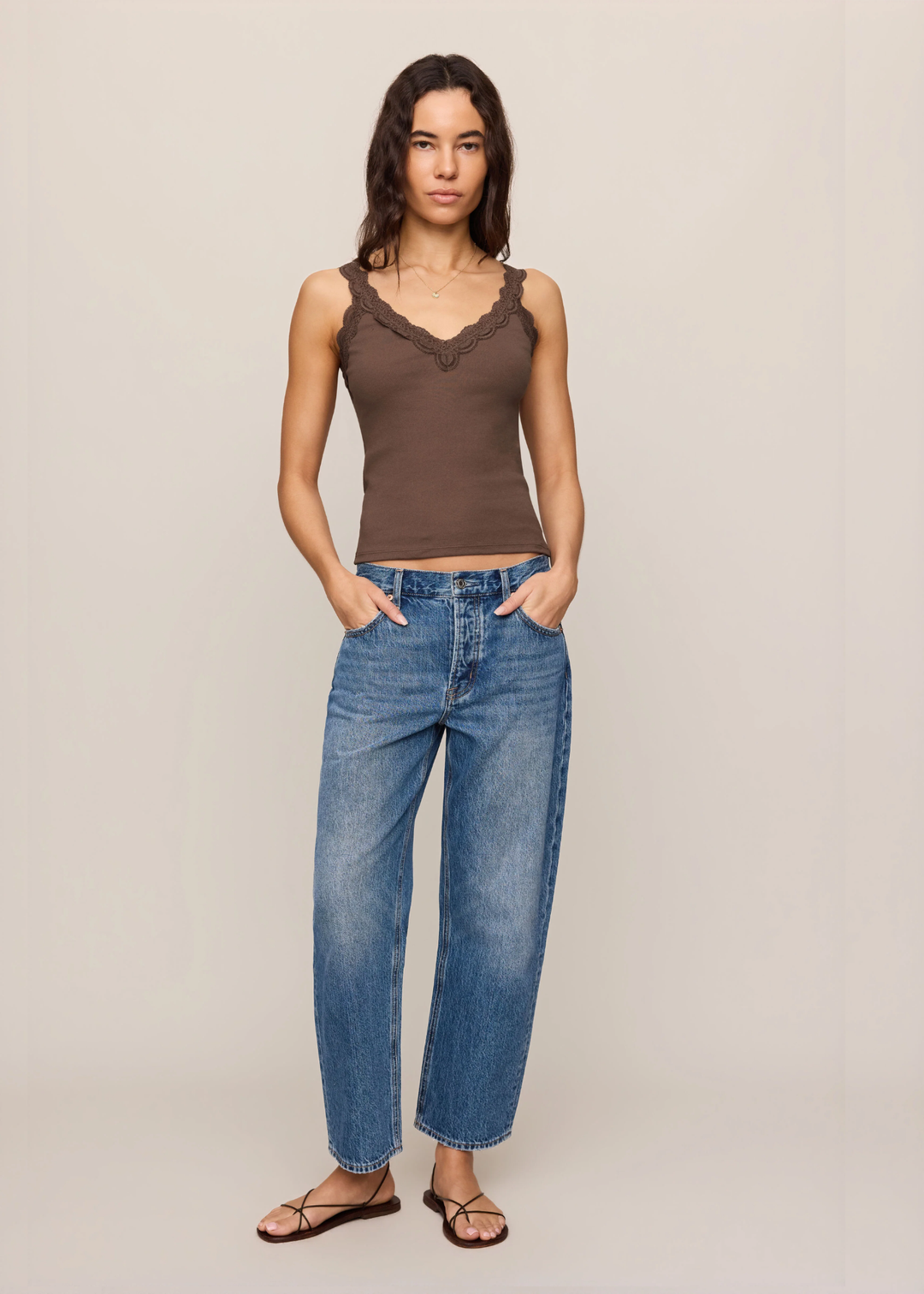 A woman modeling a brown lace-trimmed sleeveless top and blue wide-leg jeans against a plain beige studio background.