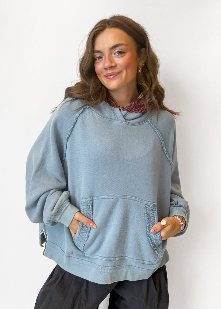 A woman wearing a light blue oversized pullover hoodie with a kangaroo pocket, layered over a red patterned shirt and paired with dark pants, standing against a plain white wall.
