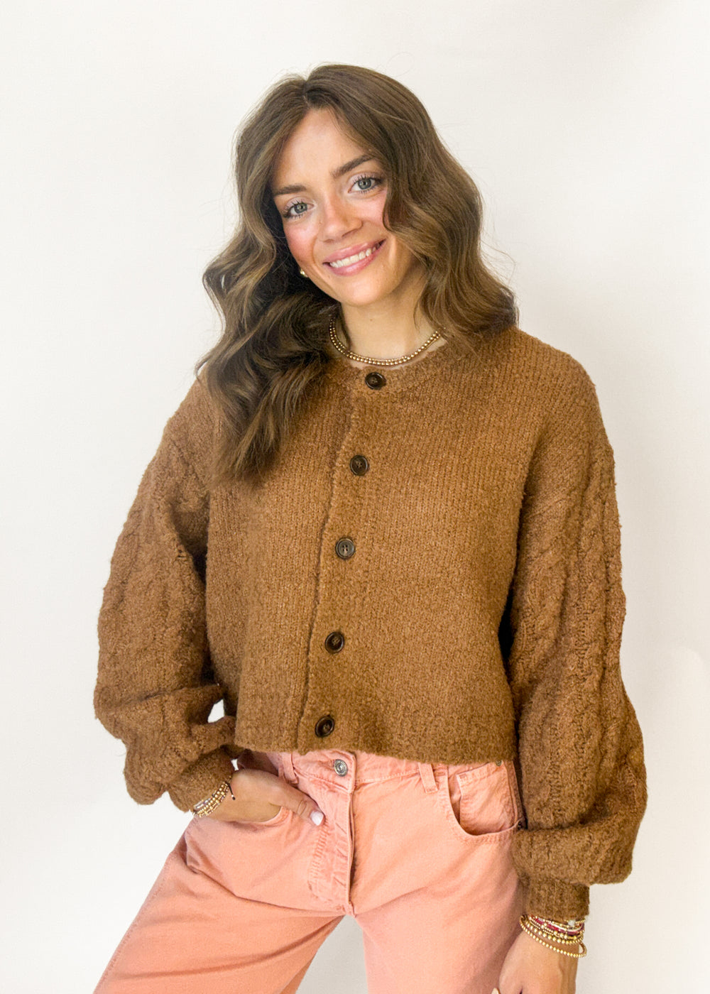 A woman wearing a brown chunky knit button-up cardigan and light pink pants, standing against a plain white background.