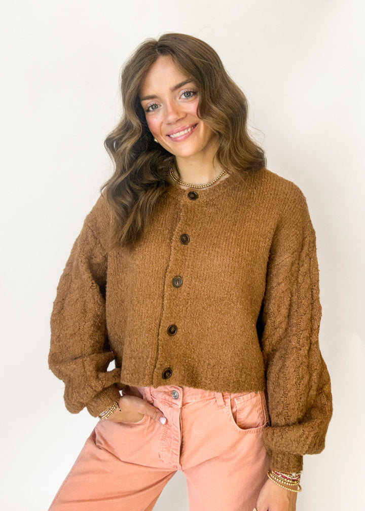 A woman wearing a brown chunky knit button-up cardigan and light pink pants, standing against a plain white background.