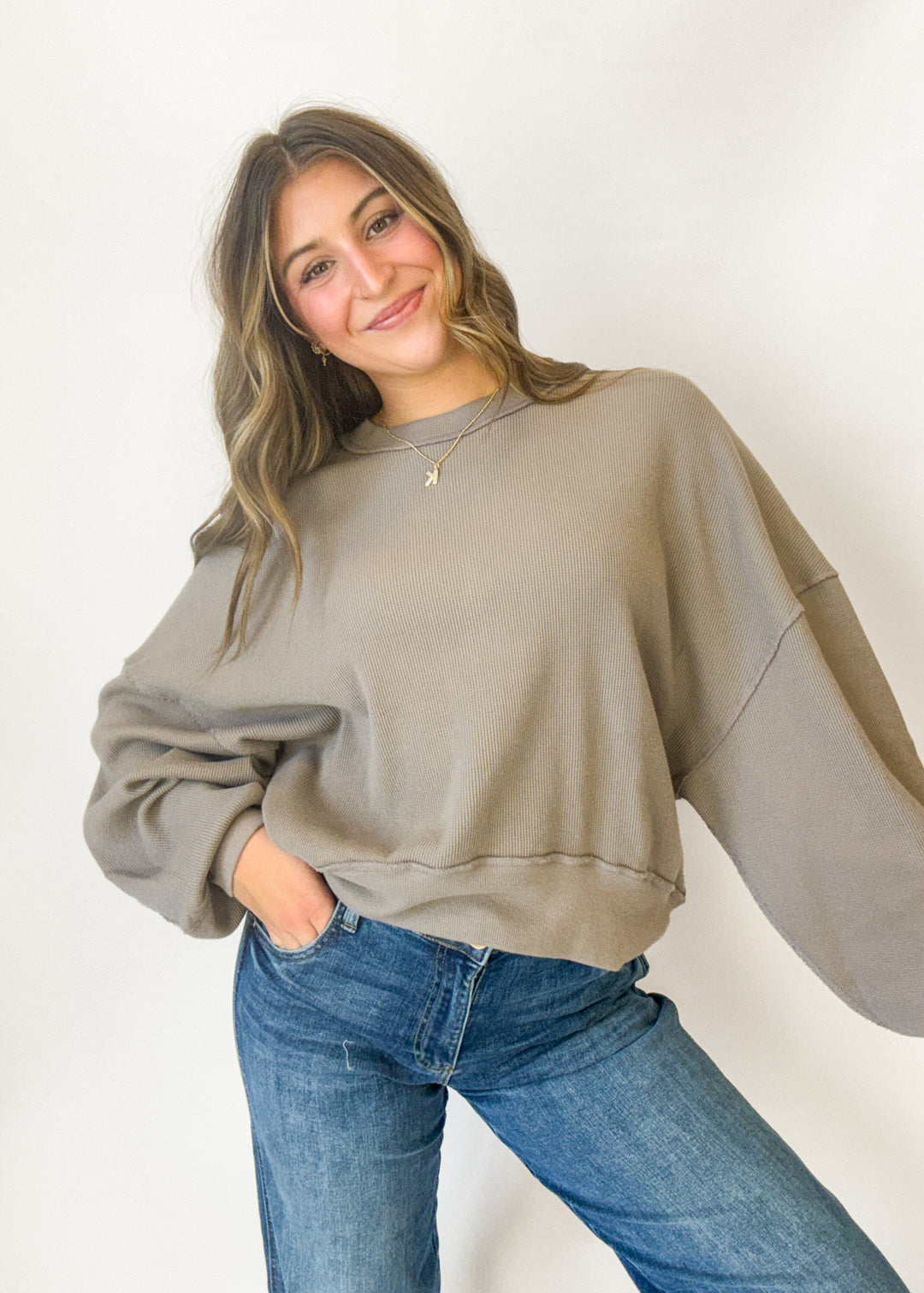 A woman wearing a taupe oversized crewneck sweatshirt and blue jeans, posing against a plain white studio background.