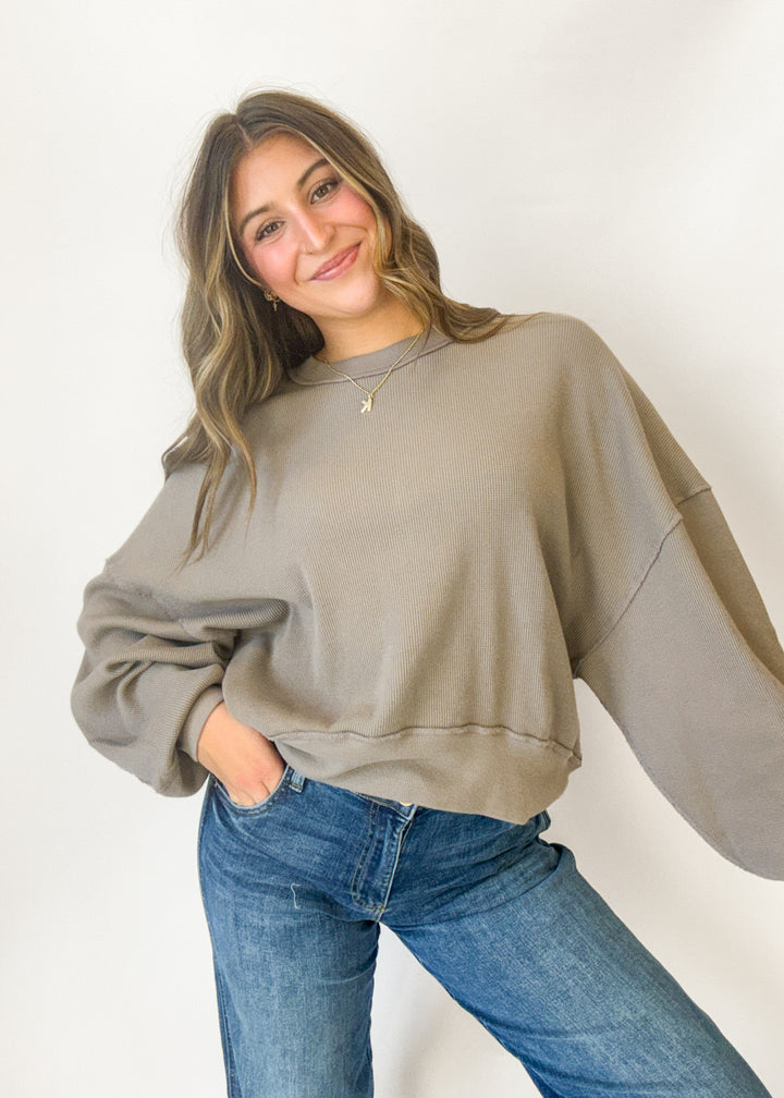A woman wearing a taupe oversized crewneck sweatshirt and blue jeans, posing against a plain white studio background.