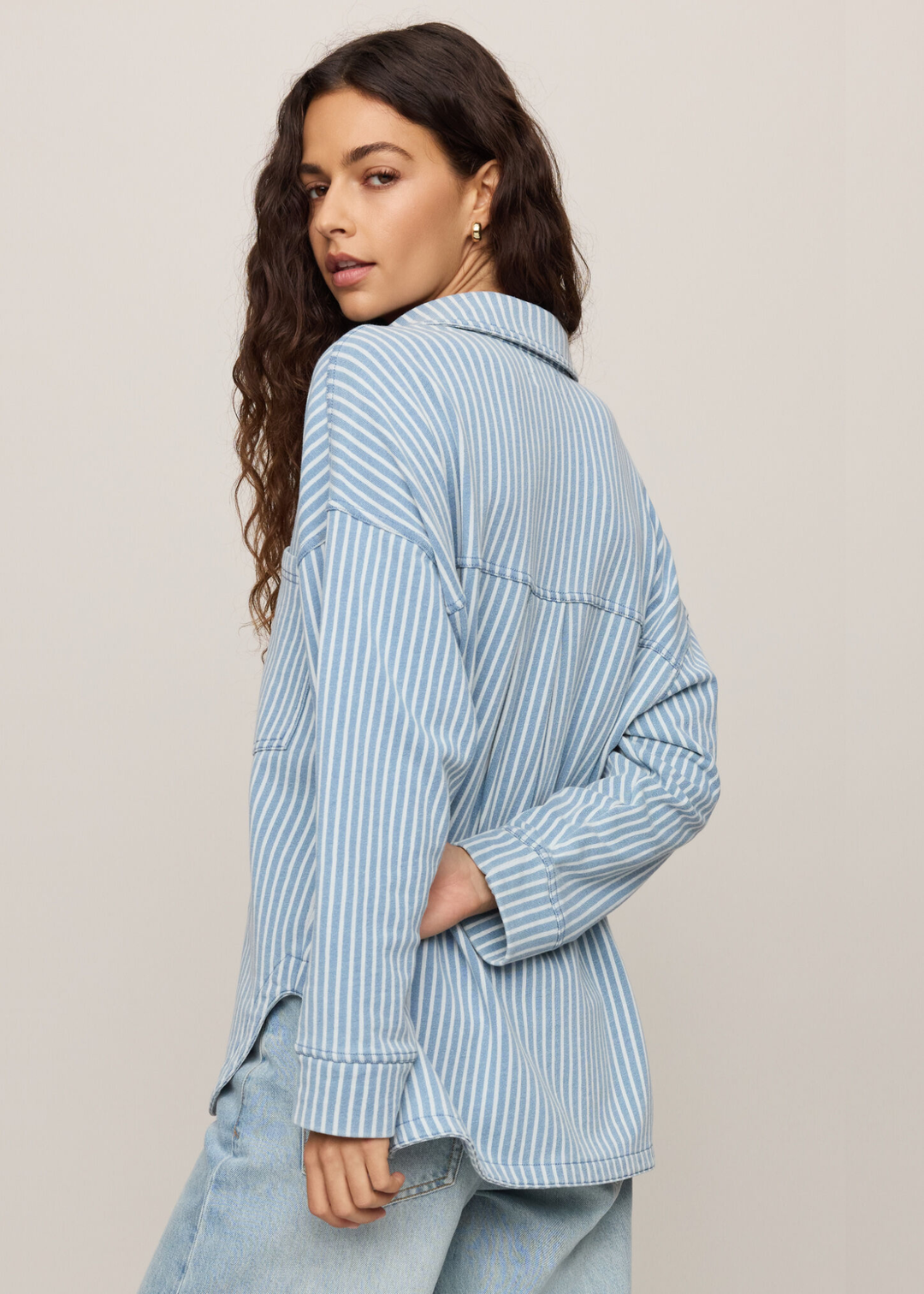 Woman wearing denim with white striped button down, turned around looking over her shoulder. 